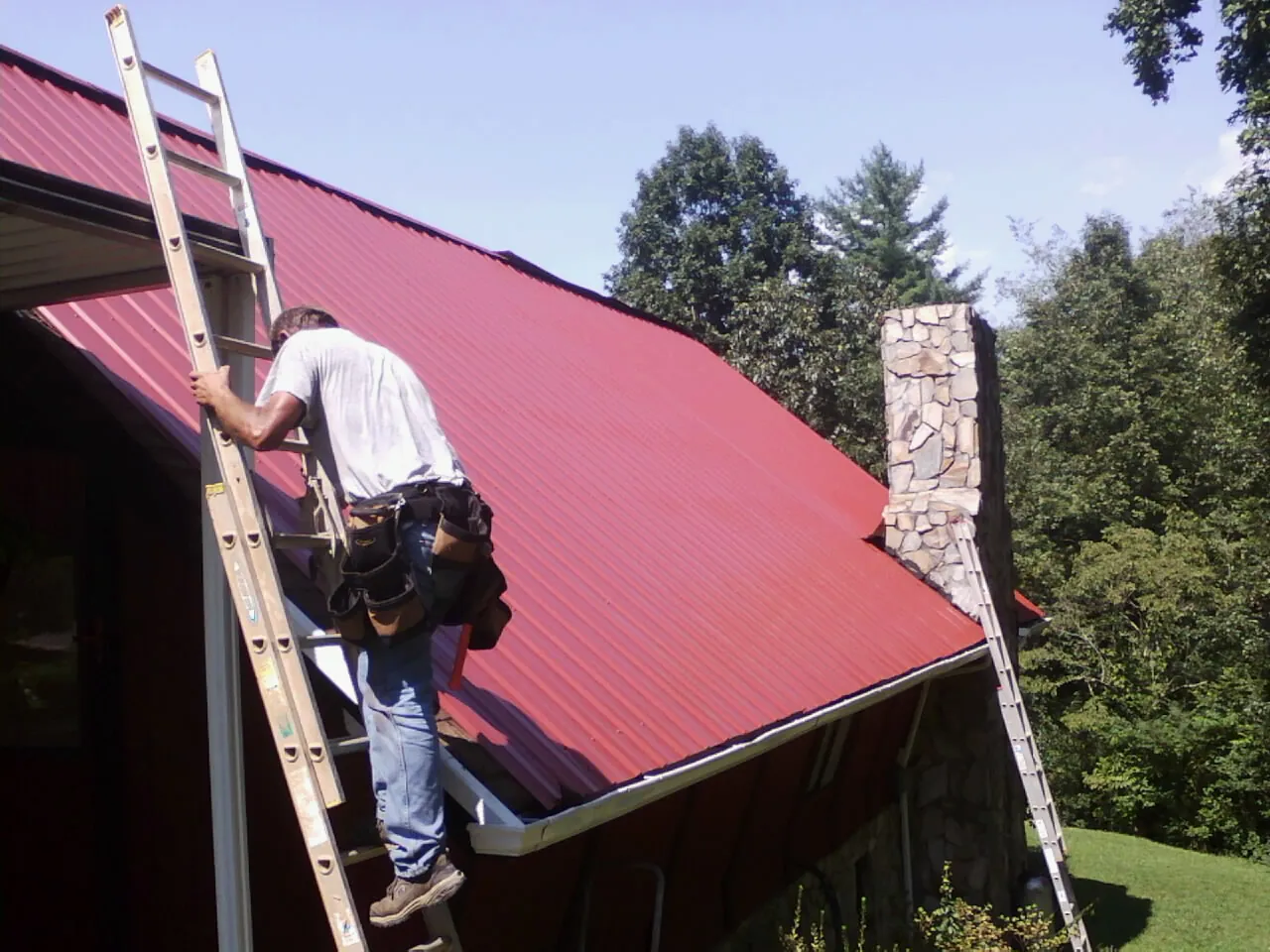Roofer on a ladder beside a red metal roof and stone chimney — Soffit & Fascia Repair by Roofing Brothers in Halethorpe
